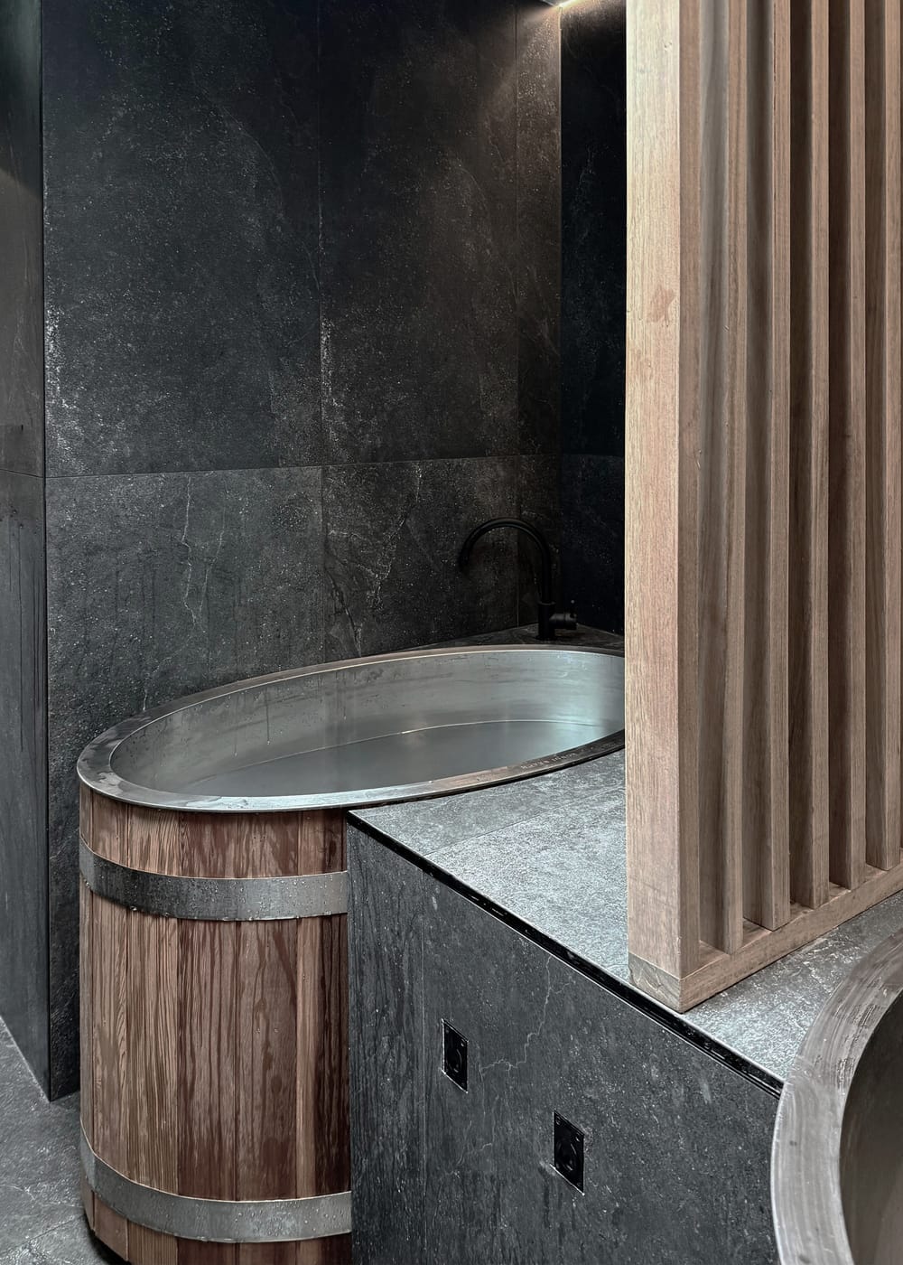 A timber barrel cold plunge with a steel liner beside dark stone